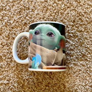 May include: White ceramic mug featuring a full-color image of Grogu, also known as Baby Yoda, from The Mandalorian. The mug has a white handle and a glossy finish. The image shows Grogu in his tan cloak, holding a blue object.
