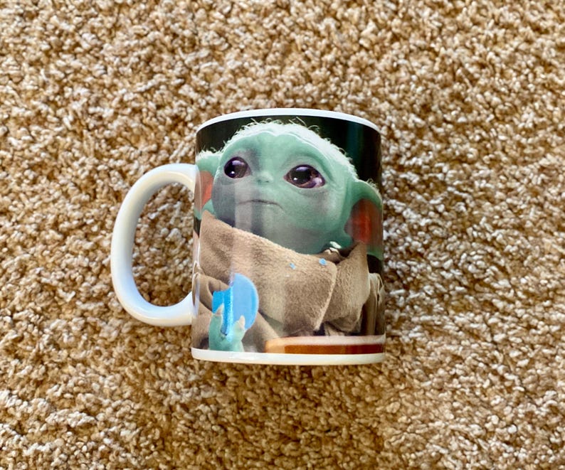 May include: White ceramic mug featuring a full-color image of Grogu, also known as Baby Yoda, from The Mandalorian. The mug has a white handle and a glossy finish. The image shows Grogu in his tan cloak, holding a blue object.