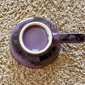 以下が含まれることがあります： 白い内側の縁とマッチするハンドルが付いた紫色のセラミックコーヒーマグ。マグカップは外側に黒と紫のデザインが施されています。マグカップは茶色のカーペットの上に置かれています。
