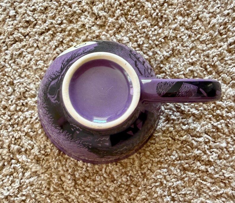 以下が含まれることがあります： 白い内側の縁とマッチするハンドルが付いた紫色のセラミックコーヒーマグ。マグカップは外側に黒と紫のデザインが施されています。マグカップは茶色のカーペットの上に置かれています。
