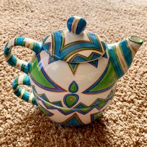 May include: A white ceramic teapot with a blue, green, and gold geometric pattern. The teapot has a lid and two handles.