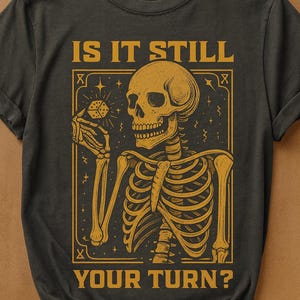 May include: Dark gray t-shirt with a yellow skeleton graphic holding a die. The text "IS IT STILL YOUR TURN?" is printed above and below the skeleton image. The design has a vintage, gothic aesthetic.