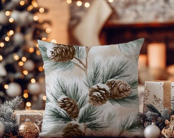 Può includere: Un cuscino quadrato con una stampa di pigne marroni e aghi di pino verdi su uno sfondo color crema chiaro. Il design presenta accenti bianchi sulle pigne e sugli aghi, che suggeriscono un leggero strato di neve depositato sopra.