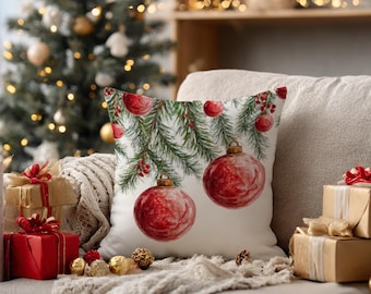 Op de afbeelding: Een vierkant sierkussen met een aquarelillustratie van groene dennentakken met rode bessen en hangende rode kerstballen tegen een witte achtergrond. Het ontwerp bevat verschillende ronde kerstballen in diverse tinten rood.