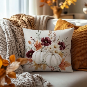 May include: A square throw pillow featuring a white background with a printed illustration of two white pumpkins surrounded by dark red flowers, cream-coloured blossoms, and various autumn-toned leaves in shades of orange, brown, and muted green.