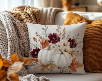 May include: A square throw pillow featuring a white background with a printed illustration of two white pumpkins surrounded by dark red flowers, cream-colored blossoms, and various autumn-toned leaves in shades of orange, brown, and muted green.
