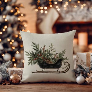 May include: A square throw pillow with a cream-colored fabric cover featuring a printed illustration of a dark green vintage sleigh filled with evergreen boughs, pine needles, red berries, and a small pinecone centered on the front surface.