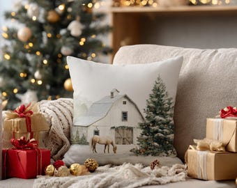 Puede incluir: Un cojín cuadrado con una ilustración impresa de un granero blanco y un caballo en un paisaje nevado con un árbol de Navidad decorado. La escena incluye colores invernales suaves y una representación detallada de un entorno rural.