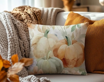 May include: A square throw pillow featuring a watercolor illustration of three pumpkins in shades of cream, orange, and sage green, accented with soft botanical leaves and flowers against a light, neutral background.
