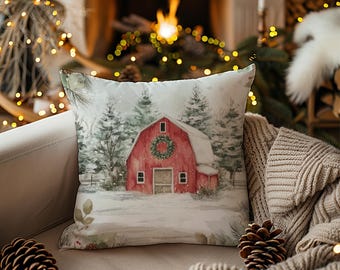 Op de afbeelding: Een vierkant sierkussen met een aquarelillustratie van een rode schuur met een groene krans op de voorgevel, omringd door besneeuwde dennenbomen en een wit winterlandschap, gedrukt op een lichtgekleurde stof met een zachte textuur.