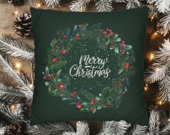 Pode incluir: Uma almofada quadrada verde-escura com uma ilustração central de uma coroa circular feita de ramos de pinheiro, folhas verdes e bagas vermelhas. O texto Merry Christmas está impresso em letra cursiva branca no centro do design.