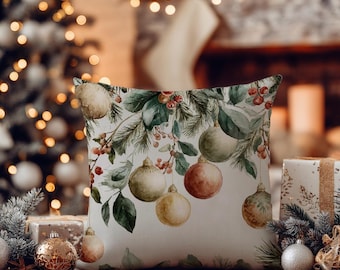 Puede incluir: Un cojín cuadrado con una ilustración en acuarela de adornos navideños colgantes, ramas de pino y bayas rojas sobre un fondo blanco. El diseño incluye esferas redondas en tonos dorados, verdes y rojos suaves, suspendidas de ramas de pino.