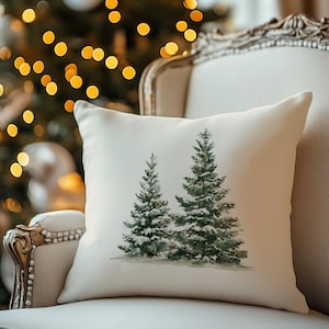May include: A square throw pillow with an off-white fabric cover featuring a centered watercolor illustration of two evergreen pine trees with white snow-dusted branches. The design is printed in shades of dark green, muted grey, and white.