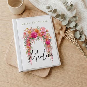 May include: A white binder with the text "MEINE ZEUGNISSE" printed in gold at the top. The cover features a colourful watercolour illustration of pink, orange, and red flowers arranged in an arch, with the name "Marlene" written in black script below.