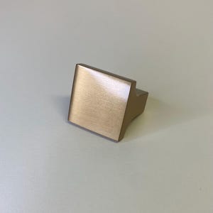 May include: A square-shaped cabinet knob with a brushed metallic finish in a warm champagne gold tone. The knob features a flat, angled front face that tapers toward the base, providing a clean, geometric profile for furniture or cabinetry hardware.