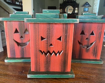 May include: Three rectangular wooden lanterns with a distressed orange and black finish and green lids and bases. Each lantern features a carved jack-o'-lantern face with triangular eyes and a jagged mouth, designed to hold a light source inside.