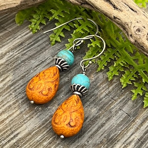 May include: A pair of handcrafted earrings featuring orange teardrop-shaped beads with intricate carvings, turquoise beads, and silver-tone hooks. The earrings are displayed against a rustic wooden surface with green foliage and a piece of driftwood.