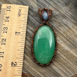 May include: A green gemstone pendant with copper wire wrapping. The pendant is oval shaped and has a light blue gemstone at the top.