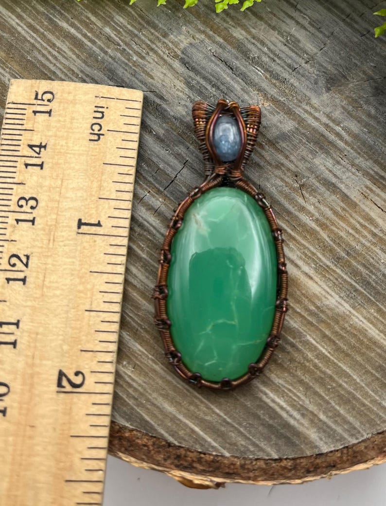 May include: A green gemstone pendant with copper wire wrapping. The pendant is oval shaped and has a light blue gemstone at the top.