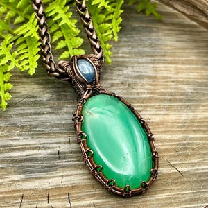 May include: A copper wire wrapped pendant featuring a green oval gemstone with a blue gemstone accent. The pendant is hanging from a copper chain.