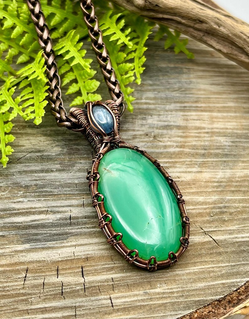 May include: A copper wire wrapped pendant featuring a green oval gemstone with a blue gemstone accent. The pendant is hanging from a copper chain.