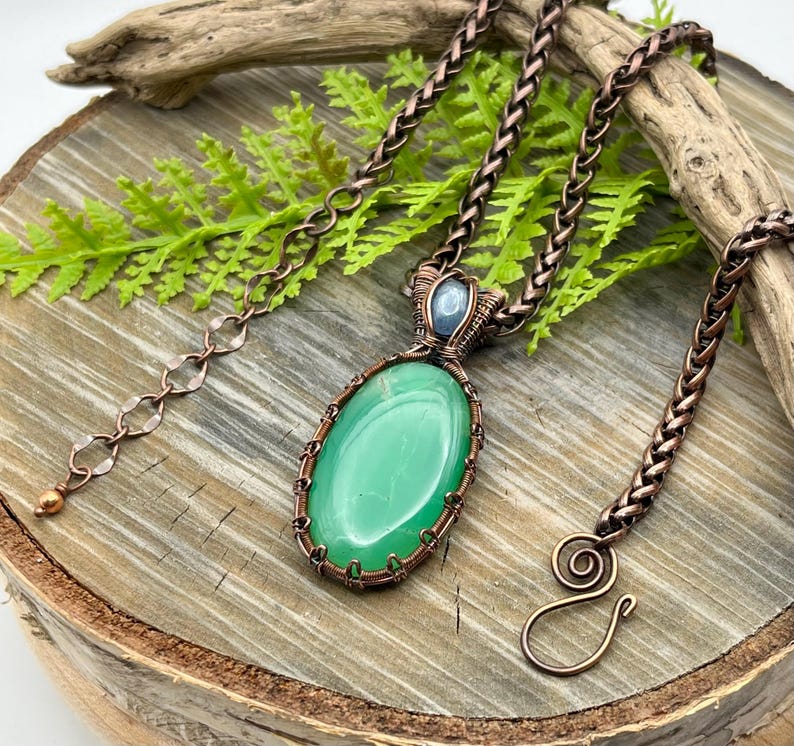 May include: A copper wire wrapped pendant necklace featuring a green oval gemstone and a smaller blue gemstone. The necklace has a copper chain with a lobster clasp.