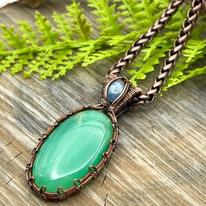 May include: A green gemstone pendant with copper wire wrapping, accented with a smaller blue gemstone. The pendant is attached to a copper chain.