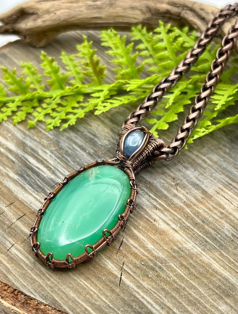 May include: A green gemstone pendant with copper wire wrapping, accented with a smaller blue gemstone. The pendant is attached to a copper chain.