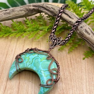 May include: A crescent moon pendant necklace. The pendant features a turquoise stone wrapped in copper wire, suspended from a copper chain. The necklace is displayed on a wooden surface with greenery in the background.