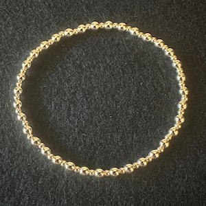 May include: A gold beaded bracelet. The bracelet is made of small, round, gold-colored beads strung together to form a circular shape. The beads have a shiny, reflective surface. The bracelet is displayed on a dark background.