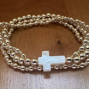 May include: Gold beaded bracelet with a white cross charm. The bracelet features multiple strands of gold-colored beads and a pearlescent white cross at the center. The bracelet is displayed on a wooden surface.