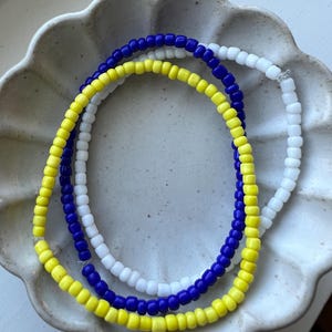 May include: A close-up of a bracelet made with yellow, white, and blue beads. The bracelet is on a white ceramic plate with a scalloped edge.