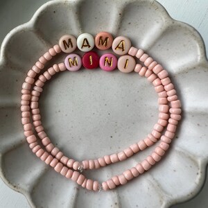 May include: Two pink beaded bracelets spelling "MAMA" and "MINI" in gold lettering on colourful beads. The bracelets are presented on a white, scalloped-edge plate.