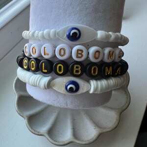 May include: A set of four bracelets with black and white beads and gold lettering spelling "COLOMA". The bracelets are stacked on top of each other and feature a blue evil eye charm.