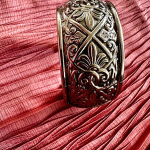 Pode incluir: Pulseira de punho prateada ornamentada com intrincados desenhos florais e geométricos. A pulseira larga tem uma forma ligeiramente curva. O fundo é um tecido texturizado rosa-avermelhado.