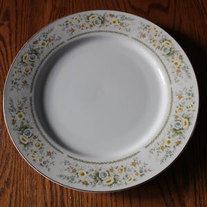 May include: A white ceramic dinner plate with a floral design in shades of yellow, blue, and green. The plate has a gold rim.