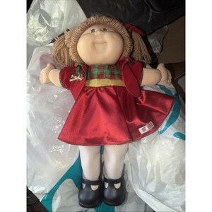 Puede incluir: Una muñeca con cabello de lana castaño claro en coletas, ojos verdes y cara redonda. Viste un vestido de satén rojo con corpiño de cuadros verdes y rojos, una cintura dorada y un pequeño lazo dorado. Lleva medias blancas y zapatos negros.