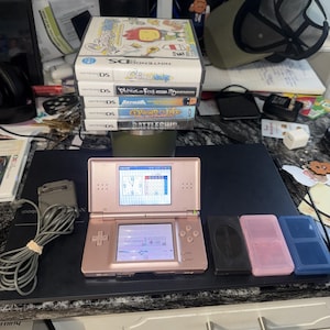 May include: A rose gold Nintendo DS handheld gaming console with a dual-screen display, a grey power adapter, and a stack of five Nintendo DS game cases including Scribblenauts, Phineas and Ferb, Batman, Drawn to Life, and Battleship, plus three game cartridges.