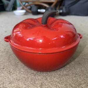 Puede incluir: Una olla de hierro fundido esmaltado de color rojo brillante con forma de manzana y tapa a juego. La tapa presenta un asa en forma de tallo de color marrón oscuro y un diseño redondeado y segmentado. La olla tiene asas laterales pequeñas.
