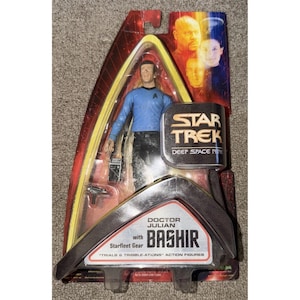 May include: A Star Trek Deep Space Nine action figure of Doctor Julian Bashir in original packaging. The figure is wearing a blue uniform and holding a device. The packaging features the Star Trek logo and images of other characters.