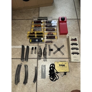 May include: A collection of model train components, including various train cars in clear plastic cases, track pieces, a red control box, and a TYCO transformer. The set includes straight and curved tracks, switches, and a cross track.