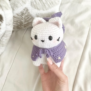 May include: White crocheted cat wearing a purple crocheted cape with white stars. The cat has black eyes and a black nose.
