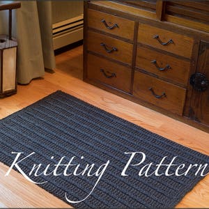 Puede incluir: Una alfombra de punto gris oscuro con textura acanalada. La alfombra está colocada sobre un suelo de madera delante de un armario de madera oscura con cajones. El texto "Knitting Pattern" está superpuesto a la alfombra.