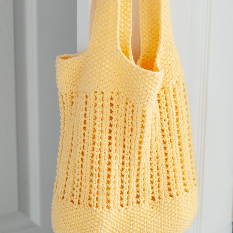May include: A yellow crocheted tote bag with a wide, open top and a woven design. The bag is hanging from a hook.