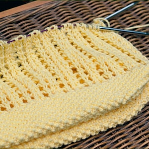 May include: A yellow knitted fabric with a lacy pattern. The fabric is folded in half and is sitting on a woven wicker surface.