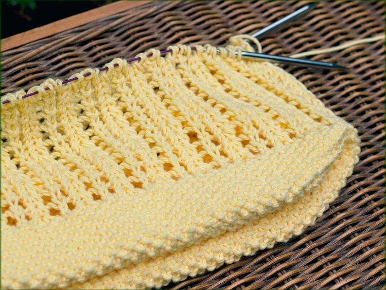 May include: A yellow knitted fabric with a lacy pattern. The fabric is folded in half and is sitting on a woven wicker surface.