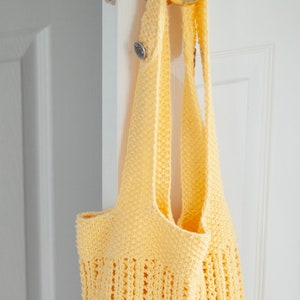 May include: A yellow crocheted tote bag hanging on a white door handle. The bag has a simple, open weave design.