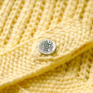 May include: A close-up of a yellow crocheted fabric with a silver button featuring a floral design.