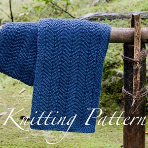 Könnte beinhalten: Ein dunkelblauer Strickschal mit strukturiertem Fischgrätenmuster, der über einen horizontalen Holzpfosten drapiert ist. Das Bild enthält den weißen Text Knitting Pattern, der unten zentriert und deutlich vor dem blauen Stoff und dem grünen Hintergrund zu sehen ist.