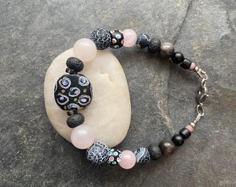 May include: A black and pink beaded bracelet with a patterned black bead, rose quartz beads, and black lava rock beads. The bracelet has a silver clasp.